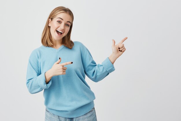 Concept publicitaire. Heureuse jeune femme européenne avec des cheveux blonds et des vêtements bleus, debout contre un mur de béton gris avec copie espace pour votre information ou contenu promotionnel