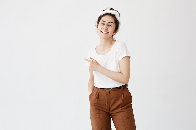 Concept publicitaire. Tir intérieur de joyeuse jeune femme souriante aux cheveux bouclés en chignon pointant l'index à l'espace de copie sur le mur blanc vierge