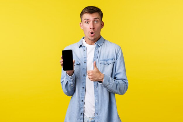 Concept de publicité de loisirs, de technologie et d'application. Un bel homme excité et surpris montrant le pouce levé comme décrit une application de téléphone portable incroyable, montrant l'écran du smartphone.