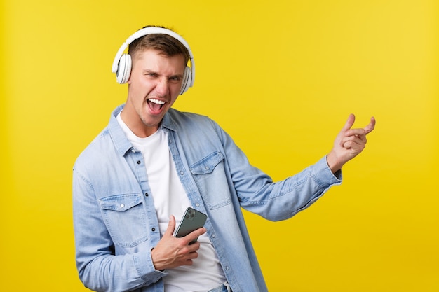 Concept de publicité de loisirs, de technologie et d'application. Un homme blond heureux et excité appréciant de la musique géniale, écoutant des chansons dans des écouteurs, tenant un téléphone portable et jouant sur une guitare invisible.