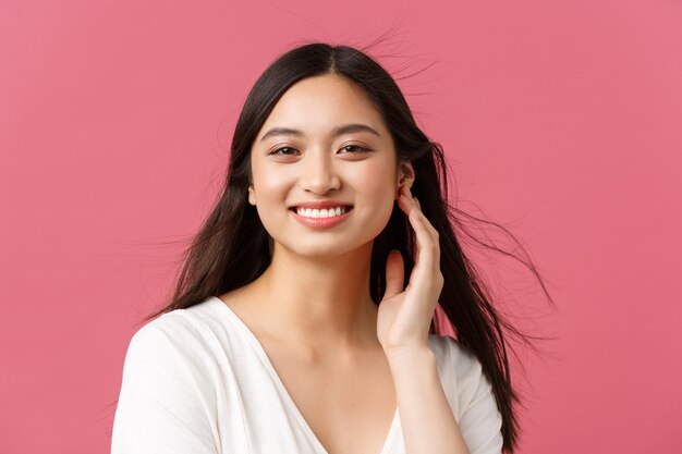 Concept de publicité pour les salons de beauté, les soins capillaires et les soins de la peau. Gros plan sur une belle jeune femme asiatique souriante alors que le vent souffle doucement sur la coupe de cheveux, debout sur fond rose.