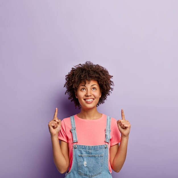 Concept de publicité et de promotion. Belle femme aux cheveux bouclés concentrée ci-dessus, pointe les deux index sur l'espace de copie, montre la direction vers le haut, porte une tenue élégante, isolée sur un mur violet