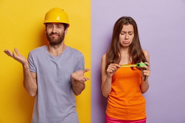 Concept de rénovation domiciliaire. Une femme triste insatisfaite regarde un ruban à mesurer, aide son mari à rénover sa maison, l'homme hésitant se tient confus, répand les paumes, porte un casque jaune, un t-shirt violet