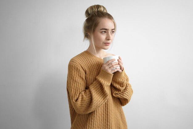 Concept de repos et de détente. Belle jeune femme portant un pull surdimensionné gardant les yeux fermés et tenant une grande tasse à deux mains, buvant du chocolat chaud ou du café à l'intérieur, souriant joyeusement