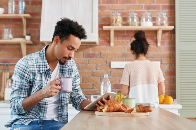 Concept de routine familiale. Mâle de race mixte lit attentivement les nouvelles sur ordinateur tablette, s'assoit à la cuisine,