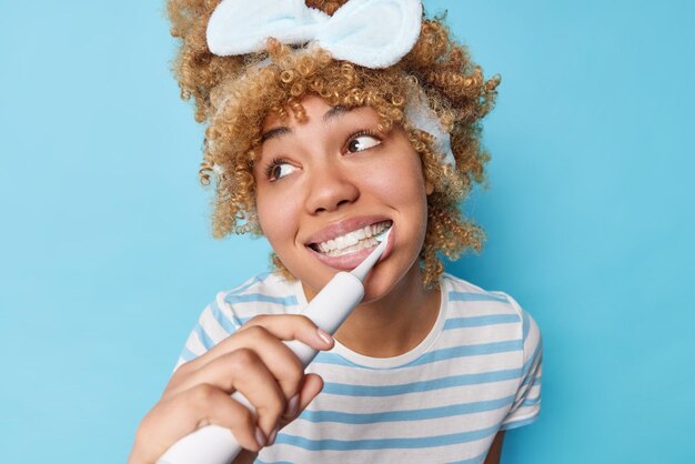 Concept de routine d'hygiène bucco-dentaire Jolie femme aux cheveux bouclés peignés regarde loin se brosse les dents avec une brosse à dents électrique rend les soins dentaires quotidiens vêtus d'un t-shirt rayé isolé sur fond bleu