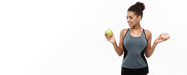 Photo gratuite concept sain et diététique belle sportive afro-américaine prendre une décision entre le beignet et la pomme verte isolé sur fond blanc