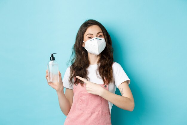 Concept de santé, de coronavirus et de distanciation sociale. Jeune femme au masque facial, portant un respirateur et pointant un désinfectant pour les mains, recommandant un antiseptique, fond bleu