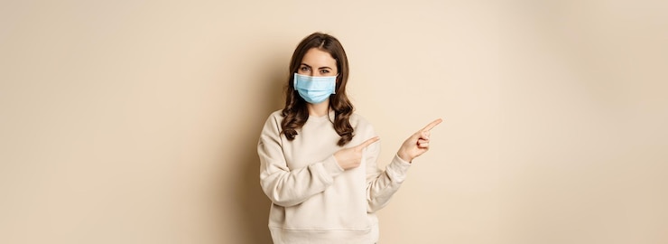 Photo gratuit concept de santé et de covid-19 une jolie femme brune portant un masque médical montrant les doigts à gauche sur la bannière du logo montrant l'espace de copie debout sur un fond beige