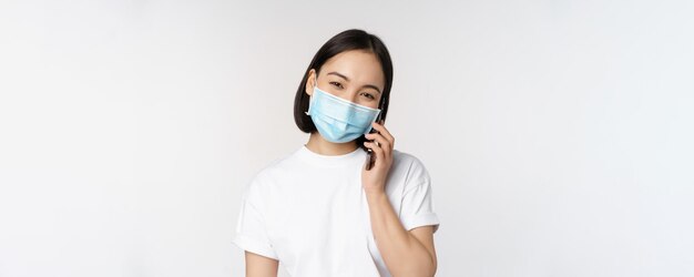 Concept de santé et de covid19 Une fille asiatique souriante dans un masque médical parlant au téléphone répond à un appel mobile debout sur fond blanc