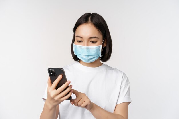 Concept de santé et de covid19 Jeune femme asiatique portant un masque médical à l'aide d'un téléphone portable en tapant sur un smartphone debout sur fond blanc