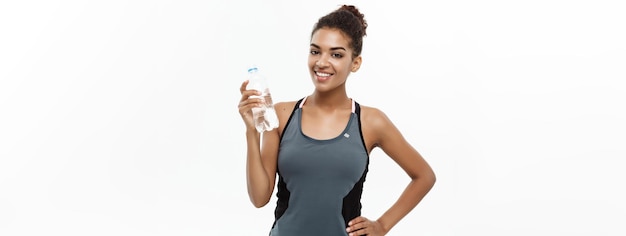 Concept de santé et de remise en forme belle fille afro-américaine en vêtements de sport tenant une bouteille d'eau en plastique après l'entraînement isolé sur fond de studio blanc