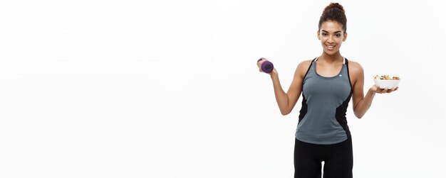 Concept de santé et de remise en forme Belle sportive afro-américaine au régime tenant des haltères et une salade fraîche sur les mains Isolé sur fond de studio blanc