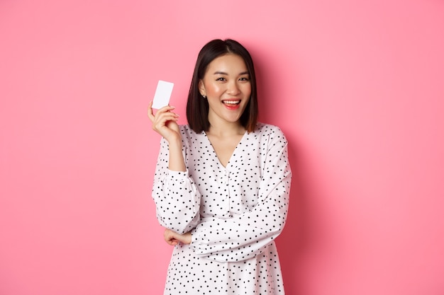 Concept de shopping femme asiatique confiante et heureuse tenant une carte de crédit et souriante satisfaite debout ...