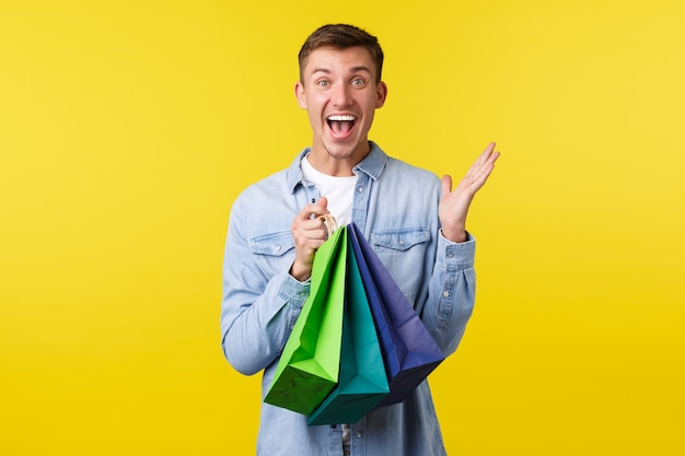 Concept de shopping, de loisirs et de réductions. Un bel homme souriant et excité crie de bonheur alors que les sacs de transport de la boutique avec des offres spéciales réagissent émerveillés par des prix merveilleux, fond jaune.