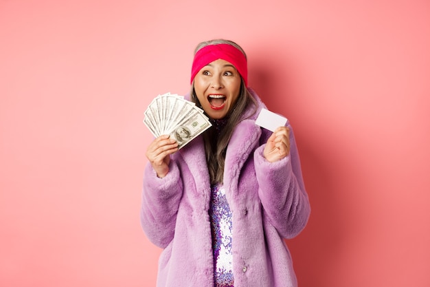 Concept de shopping et de mode. Une femme âgée asiatique crie heureuse comme un gagnant, tenant de l'argent en dollars et une carte de crédit en plastique, l'air excité, fond rose