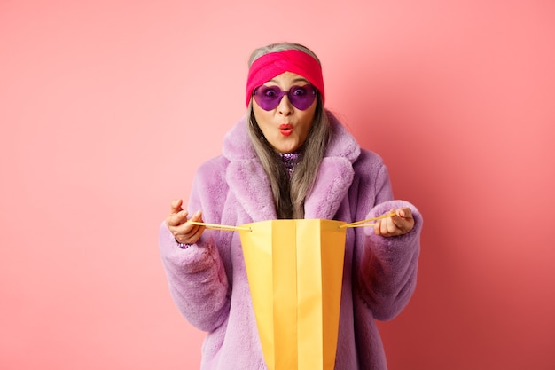 Concept de shopping et de mode. Femme âgée asiatique élégante en lunettes de soleil et manteau en fausse fourrure sac en papier ouvert avec des cadeaux, à la surprise de la caméra, fond rose