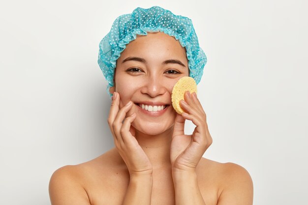 Concept de soins de la peau, de cosmétologie et de traitement du visage. Belle femme souriante applique le fond de teint sur le visage avec une éponge, a une peau lisse après avoir pris un bain, porte un bonnet de douche, des modèles sur un mur blanc