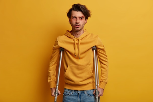 Concept de soins de santé. Un homme handicapé avec des béquilles étant handicapé après un accident tragique, a des ecchymoses et une abrasion, incapable de marcher, isolé sur un mur jaune. Aide à la mobilité. Homme blessé