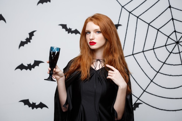 Concept de sorcière d'halloween - happy halloween witch tenant un verre de vin rouge sanglant sur un mur gris foncé avec une chauve-souris et une toile d'araignée.
