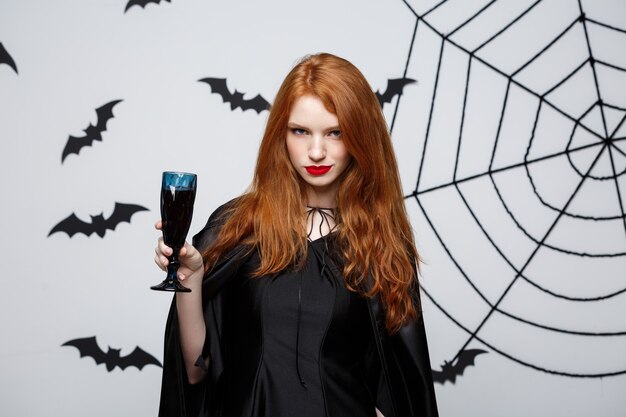Concept de sorcière d'Halloween - Happy Halloween Witch tenant un verre de vin rouge sanglant sur un mur gris foncé avec une chauve-souris et une toile d'araignée.