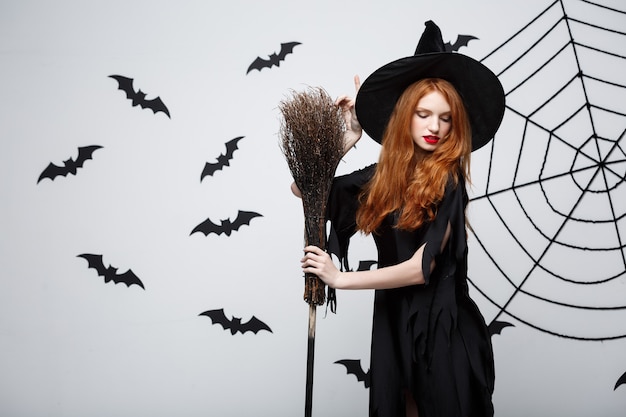 Concept de sorcière d'Halloween portrait de la belle jeune sorcière avec manche à balai sur mur gris avec mur de chauve-souris et toile d'araignée