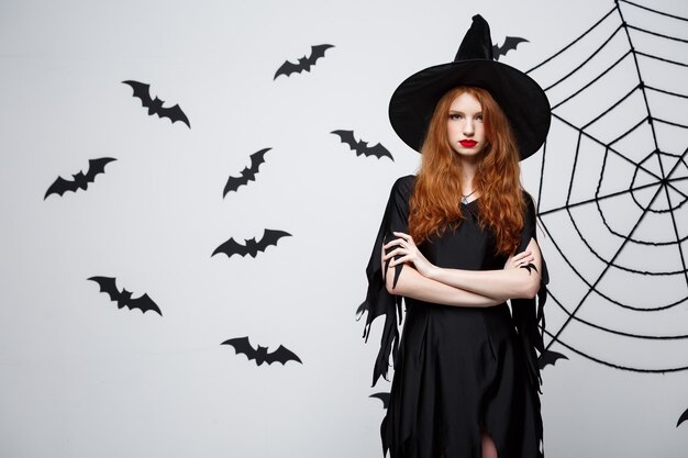 Concept de sorcière d'Halloween sorcière d'halloween tenant posant avec une expression sérieuse sur un mur gris foncé avec une chauve-souris et une toile d'araignée