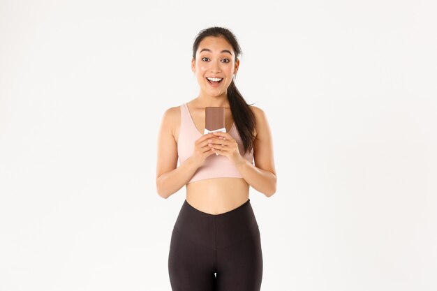 Concept de sport, bien-être et mode de vie actif. Athlète féminine asiatique souriante heureuse tenant la protéine de chocolat mauvais et à la recherche excitée, mangeant des bonbons sains pour un entraînement prolongé.