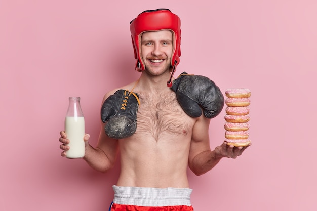 Concept de sport et de nutrition des gens. Un boxeur masculin maigre positif pose avec des sourires de torse nu a volontiers la tentation de manger des beignets et de boire du lait