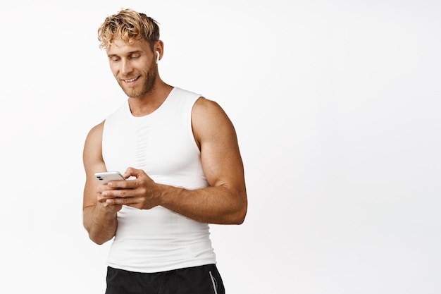 Concept de sport et de remise en forme Beau sportif masculin utilisant une application pour smartphone écoutant de la musique dans des écouteurs sans fil debout sur fond blanc