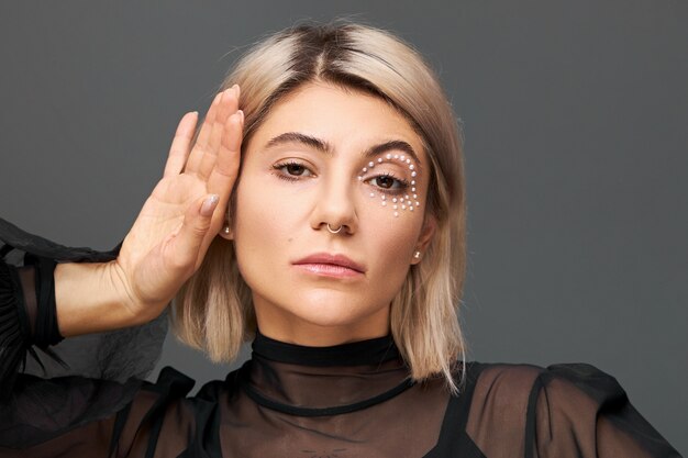 Concept de style, glamour, cosmétiques et beauté. Portrait de superbe jeune femme en chemisier transparent noir posant isolé visage touchant, portant le piercing, coupe de cheveux courte teinte et maquillage à la mode