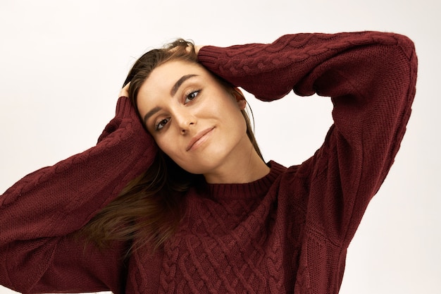 Concept de style, mode, tricot et saison. Portrait de jolie jeune femme européenne confiante habillée en pull chaud tenant les mains sur sa tête, regardant la caméra avec un sourire joyeux et joyeux