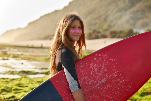 Concept de surfeur et océan. Une femme aux cheveux noirs ravie porte des looks de planche de surf cirés avec une expression satisfaite