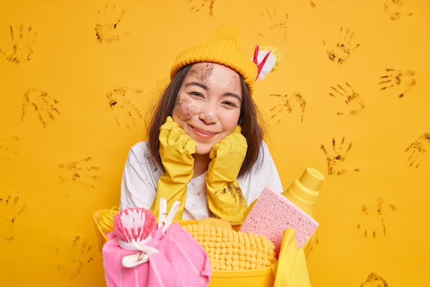 Concept de tâches ménagères Une femme asiatique heureuse se sent fatiguée mais satisfaite garde les mains sous le menton porte un chapeau et des gants en caoutchouc s'appuie sur un panier avec du linge et des détergents isolés sur fond jaune