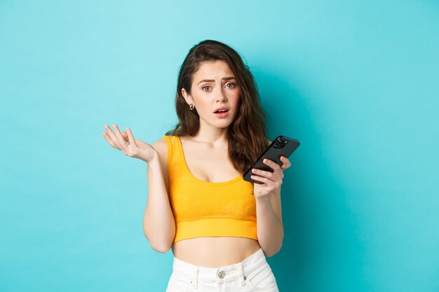 Concept de technologie et de style de vie. Une fille glamour a l'air confuse après avoir lu l'écran du smartphone, regardant la caméra perplexe, debout sur fond bleu.