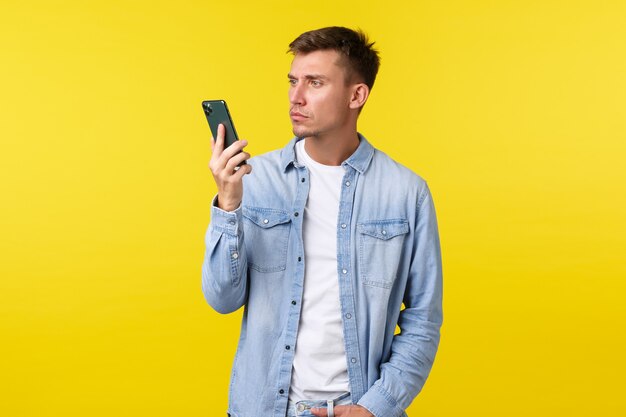 Concept de technologie, de style de vie et de publicité. Jeune homme agacé et frustré, perplexe au sujet d'une conversation soudainement terminée, regardant le téléphone portable irrité, debout sur fond jaune.