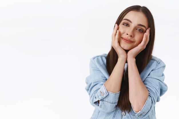 Concept de tendresse, de beauté et d'émotions. Jeune femme féminine mignonne et douce des années 20 en veste en jean, tête inclinée, joues touchantes et rêveuse souriante, regarde avec admiration et affection, fond blanc