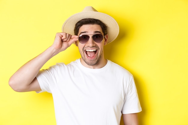 Concept de tourisme et de vacances. Gros plan d'un homme heureux en chapeau d'été et lunettes de soleil profitant de vacances, debout sur fond jaune