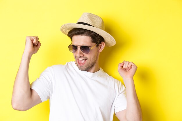 Concept de tourisme et de vacances. Gros plan d'un homme profitant de vacances en voyage, dansant et pointant du doigt sur le côté, portant des lunettes de soleil avec chapeau de paille, fond jaune.