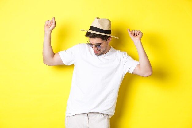 Concept de tourisme, de voyages et de vacances. Homme touristique profitant de vacances, dansant au chapeau de paille et lunettes de soleil, posant sur fond jaune