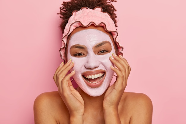 Concept de traitement du visage. Belle femme joyeuse pose avec un masque facial à l'argile, sourit largement, montre des dents blanches parfaites, porte un bandeau de douche protecteur, se soucie de sa bonne apparence, a une manucure