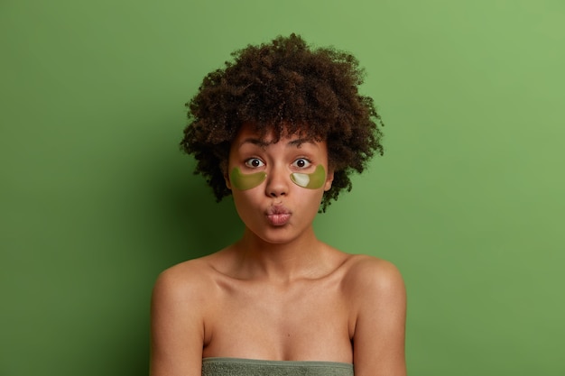 Photo gratuite concept de traitement du visage. belle jeune femme rafraîchie avec une coiffure afro, utilise des taches vertes sous les yeux, arrondit les lèvres, se tient enveloppé dans une serviette, a une beauté naturelle, pose sur un mur vert
