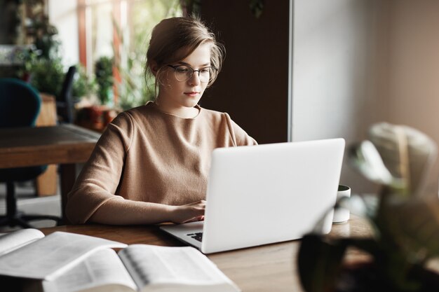 Concept de travail, de style de vie et d'entreprise. Belle femme européenne concentrée dans des verres à la mode assis dans un café près d'un ordinateur portable, travaillant sur un ordinateur portable, entouré de livres, prenant des notes.