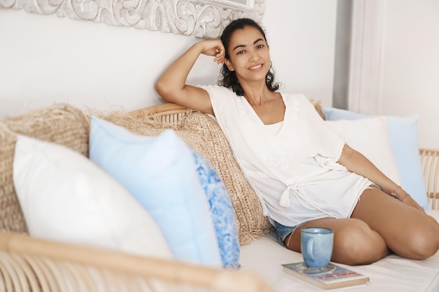 Concept de vacances bien-être bien-être Jolie femme bronzée souriante relaxante canapé boire du café frais mettre une tasse de thé livre regarder appareil photo joyeux passer les week-ends lecture paisible à la maison