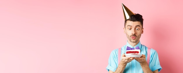 Concept de vacances et de célébration heureux jeune homme en chapeau de fête d'anniversaire soufflant une bougie sur la fabrication de gâteaux