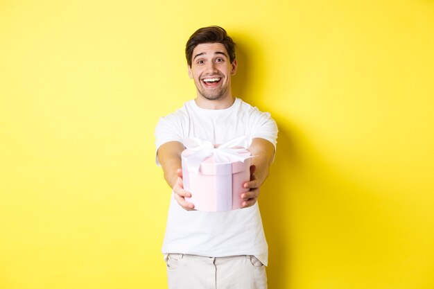 Concept de vacances et de célébration. Homme souriant vous donnant un cadeau, féliciter, debout sur fond jaune avec présent.