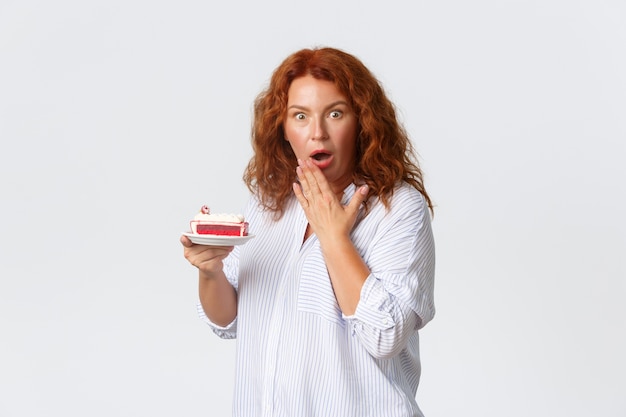 Concept de vacances, d'émotions et de style de vie. Femme rousse d'âge moyen choquée et inquiète, bouche ouverte, haletante et l'air inquiète en tenant un gâteau, entendez combien de calories il a.