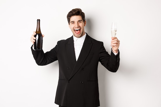 Concept de vacances, fête et célébration. Bel homme en costume tendance s'amusant, tenant une bouteille et une coupe de champagne, debout sur fond blanc.