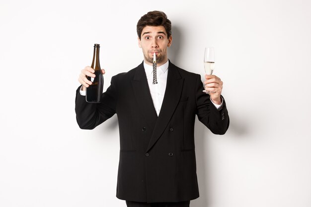 Concept de vacances, fête et célébration. Portrait de beau mec en costume noir, soulevant une bouteille de champagne et de verre, soufflant un sifflet de fête, ayant un anniversaire, debout sur fond blanc