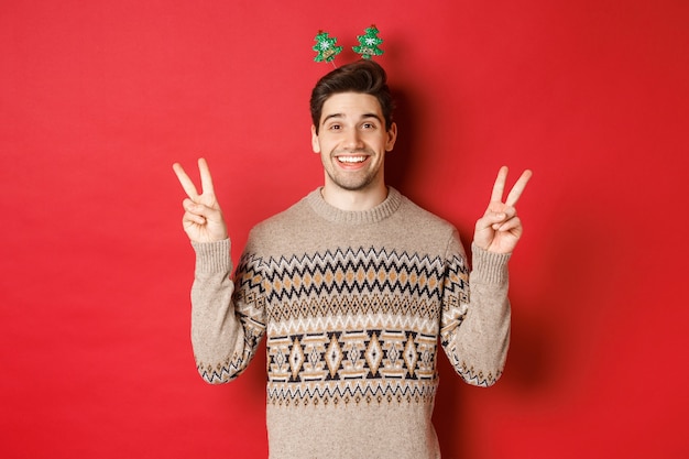 Concept de vacances d'hiver, Noël et célébration. Image d'un mec beau et idiot habillé pour la fête du nouvel an, montrant des signes de paix et souriant, debout sur fond rouge